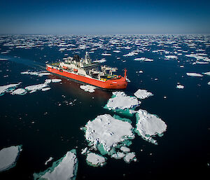 A red ship sailing between sea ice floes in a dark blue ocean.