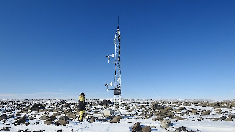 A man stands beside a weather station tower on snow covered rocks.
