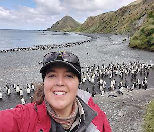 A smiling woman takes a selfie with penguins on the beach behind her.