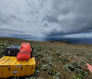 A yellow plastic box and orange bag sit on a grassy hill with grey clouds overhead