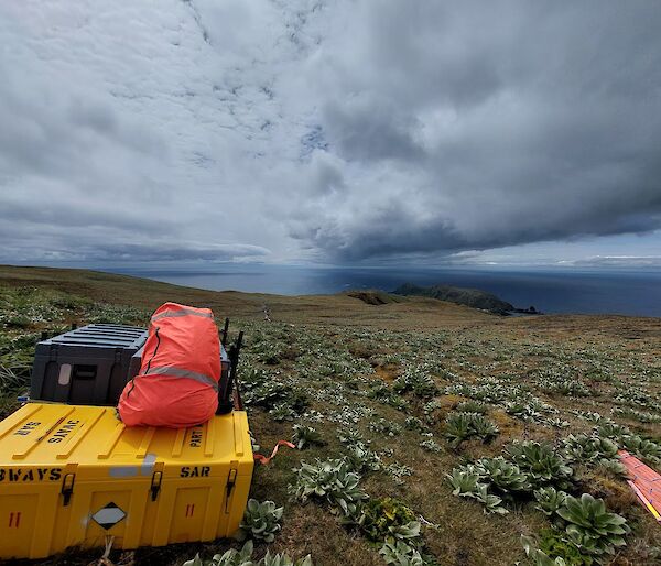 A yellow plastic box and orange bag sit on a grassy hill with grey clouds overhead