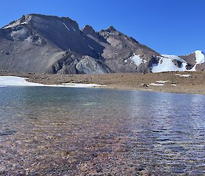 A lake in the foreground with a barren jagged hill in the background, partly covered in snow.