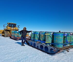 An expeditioner stands on the snow with a dozer towing a sled with fuel drums on it.