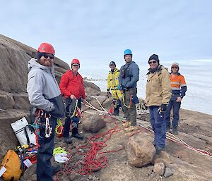 Six people in hard hats, holding ropes, stand on a rocky slope.