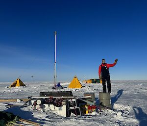 Expeditioner standing with a load of cargo in the foreground with tents and ice drill set up in the background