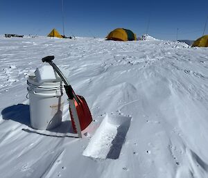 Snow shovel and white bucket on the snow in the foreground. A line of yellow tents in the background