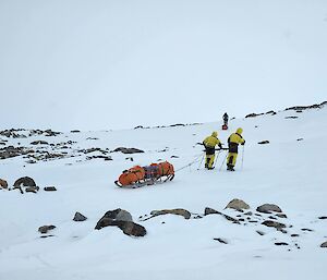 two expeditioners hauling a wooden sled up a hill in high winds and snow