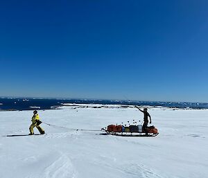 One expeditioner hauling a loaded sled with another expeditioner sitting on the back