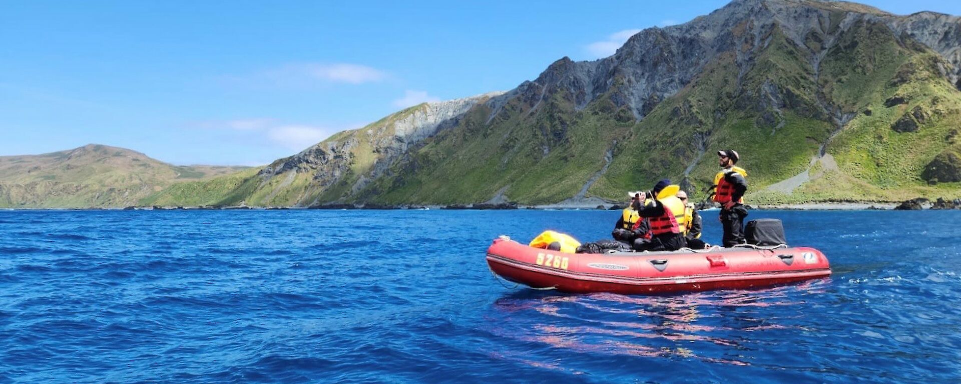 Two people in a red dinghy on blue sea with green hills behind