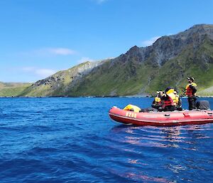 Two people in a red dinghy on blue sea with green hills behind