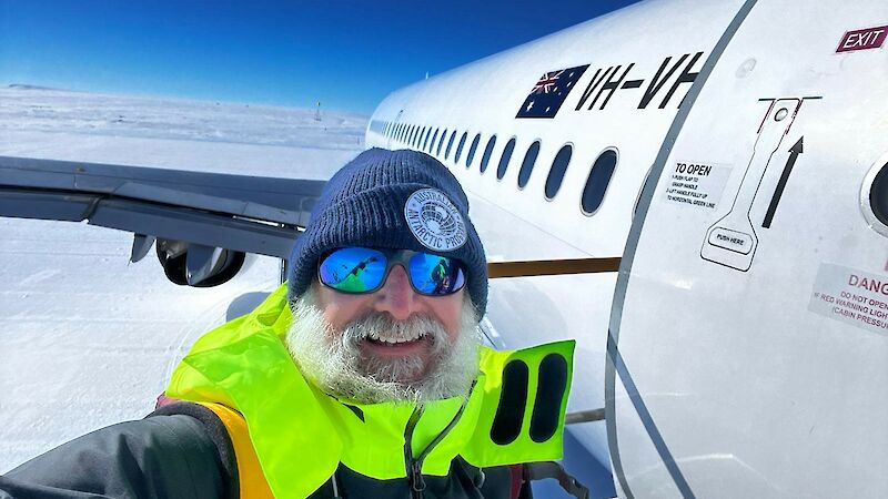 A man taking a selfie on the steps of an aircraft in Antarctica, with a view of the aircraft wing and white ice.