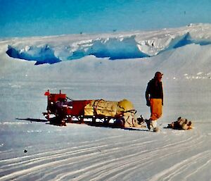 A man stands next to a sled with a large ice shelf in the background. He is wearing a brown woolly hat.
