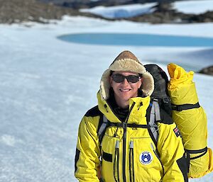 A man stands smiling with a brown woolly cap on. The background in blurred but you can see an icy landscape behind him. He is wearing a yellow suit with the Australian Antarctic Program logo.