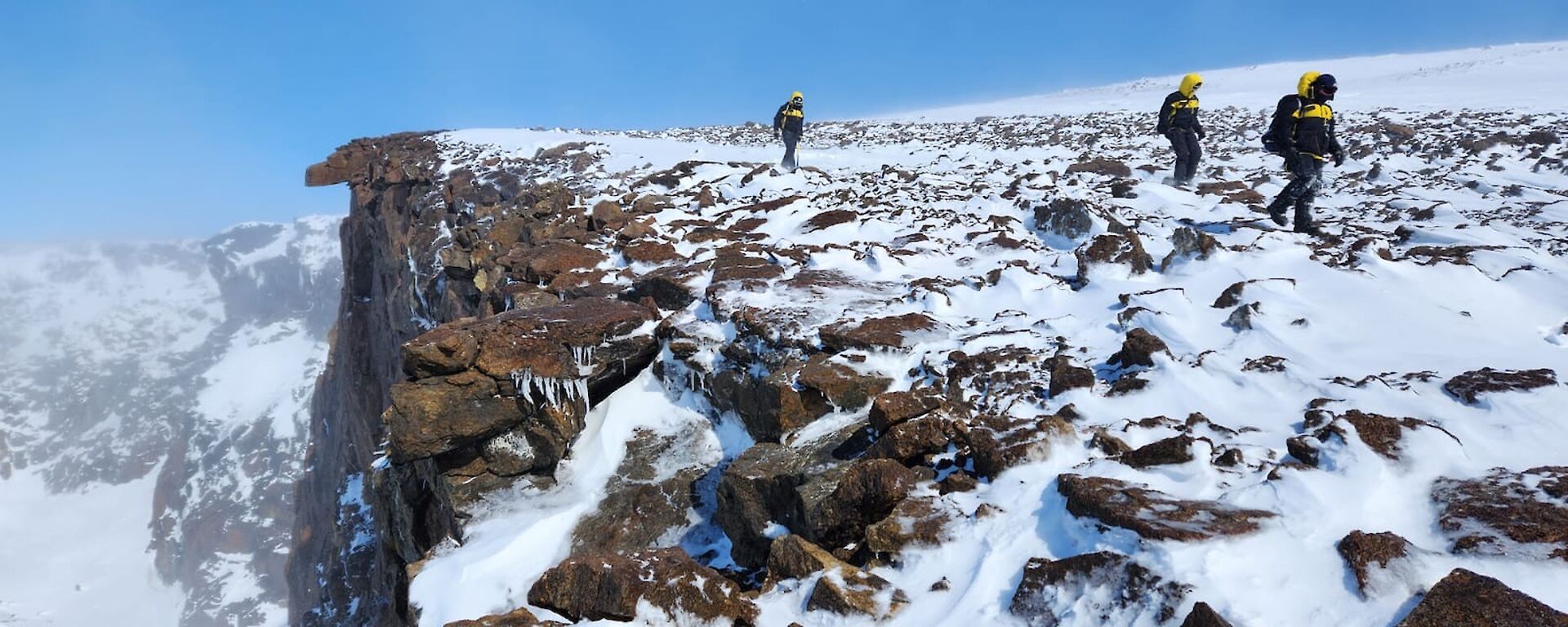 Three figures walk along a mountain ledge covered in snow