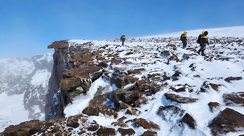 Three figures walk along a mountain ledge covered in snow