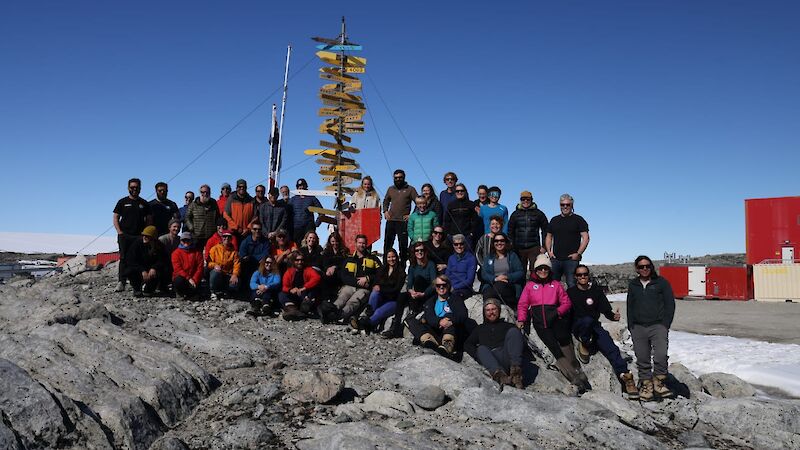 A large group stand on a rock with a signpost behind them.