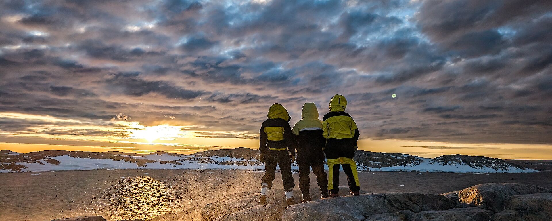 Three expeditioners standing on rocks watching the sunset on the horizon