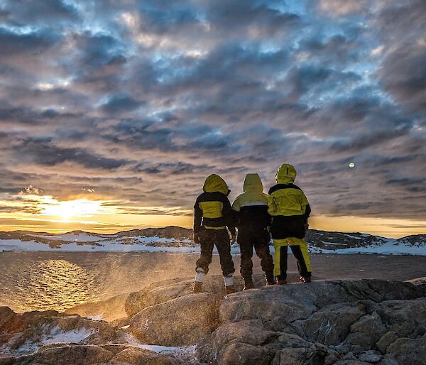 Three expeditioners standing on rocks watching the sunset on the horizon