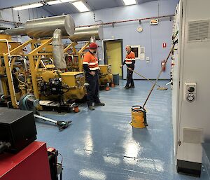 Two men with mops, cleaning up a blue room with yellow generators