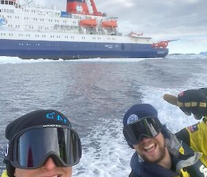 Two smiling people in snow goggle on a small boat with a large ship behind them