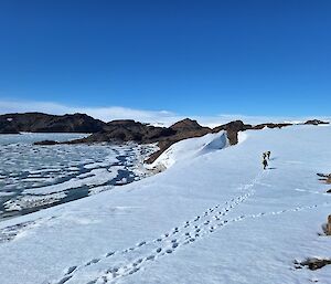 Lone figures walk up a snowy hill with ice-covered water to the left and rocky hills ahead