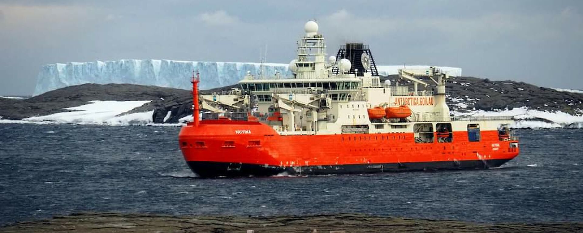 A red icebreaker ship sits in a choppy bay with a white iceberg in the background.