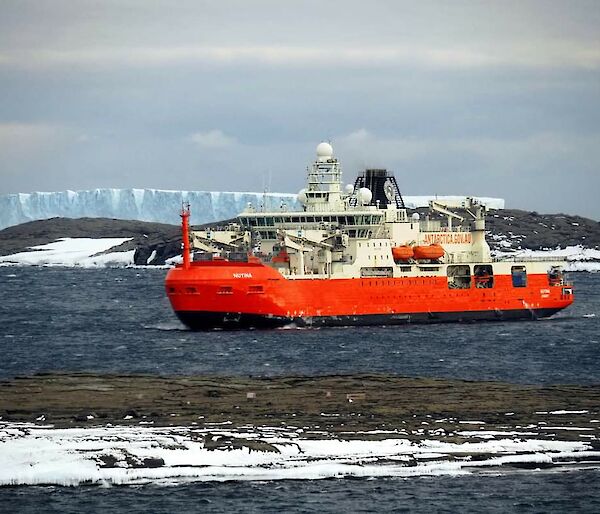 A red icebreaker ship sits in a choppy bay with a white iceberg in the background.