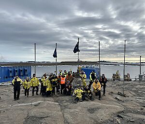 A group in yellow Antarctic clothing stand on a rocky shelf with water behind them and flags flying.