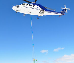 Two men stand under a helicopter with a winch, loading a crate onto it.