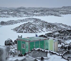 A green shed stands on a rocky hill, surrounded by snow.