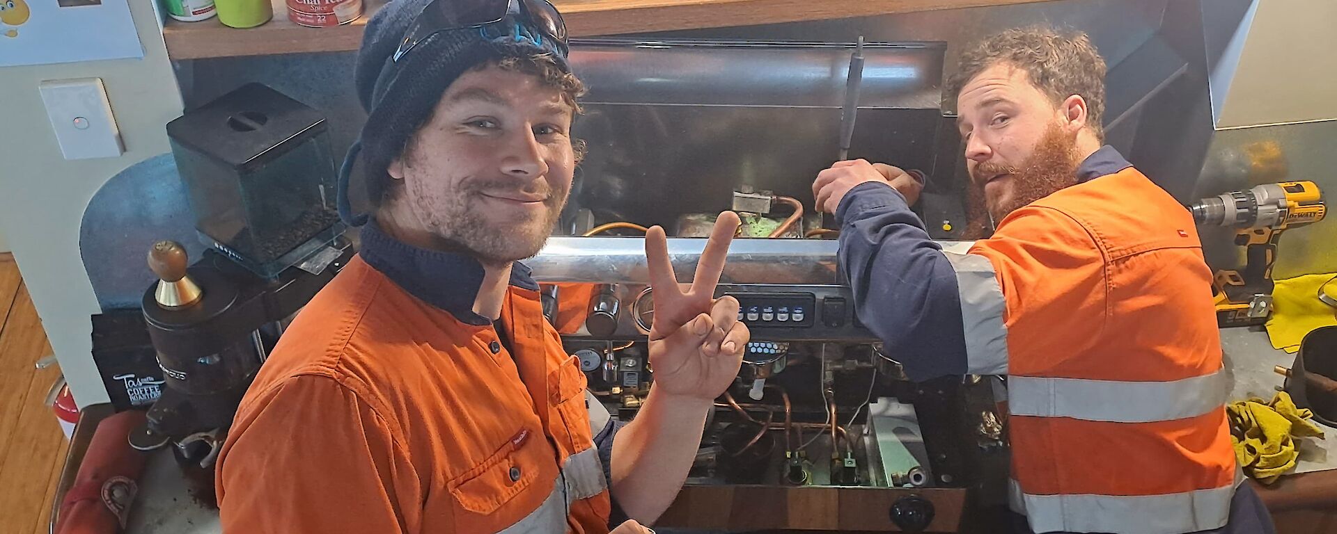 Two men in high vis work on a coffee machine.