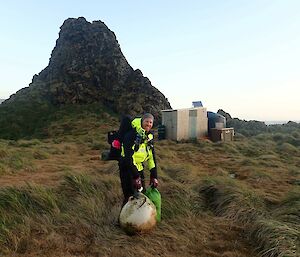 A person stands on a grassy slope at dawn carrying marine debrib