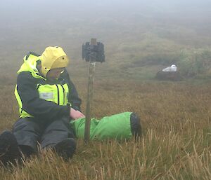 A person in yellow expeditioner gear takes something out of a bag with a camera on a tripod next to them.