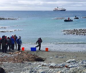 A group of people stand on a grey sand beach with a cruise ship out to sea behind them