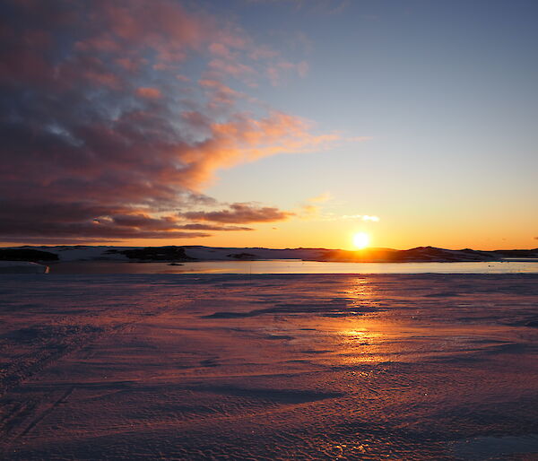 The sun rises over water and ice, with pink clouds in the sky