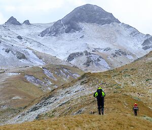 two people walk up a slope with snow-covered hills in the background