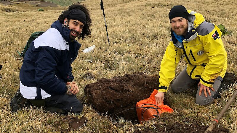 Two expeditioner place an orange box in a trench on a grassy hill.