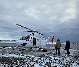 A helicopter lands at Mawson with critical cargo for station, two expeditoners standby awaiting the signal to approach from the pilot