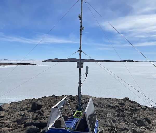 A portable weather station secured to a rock outcrop in the foreground. Sea ice and blue sky with landscape in the background