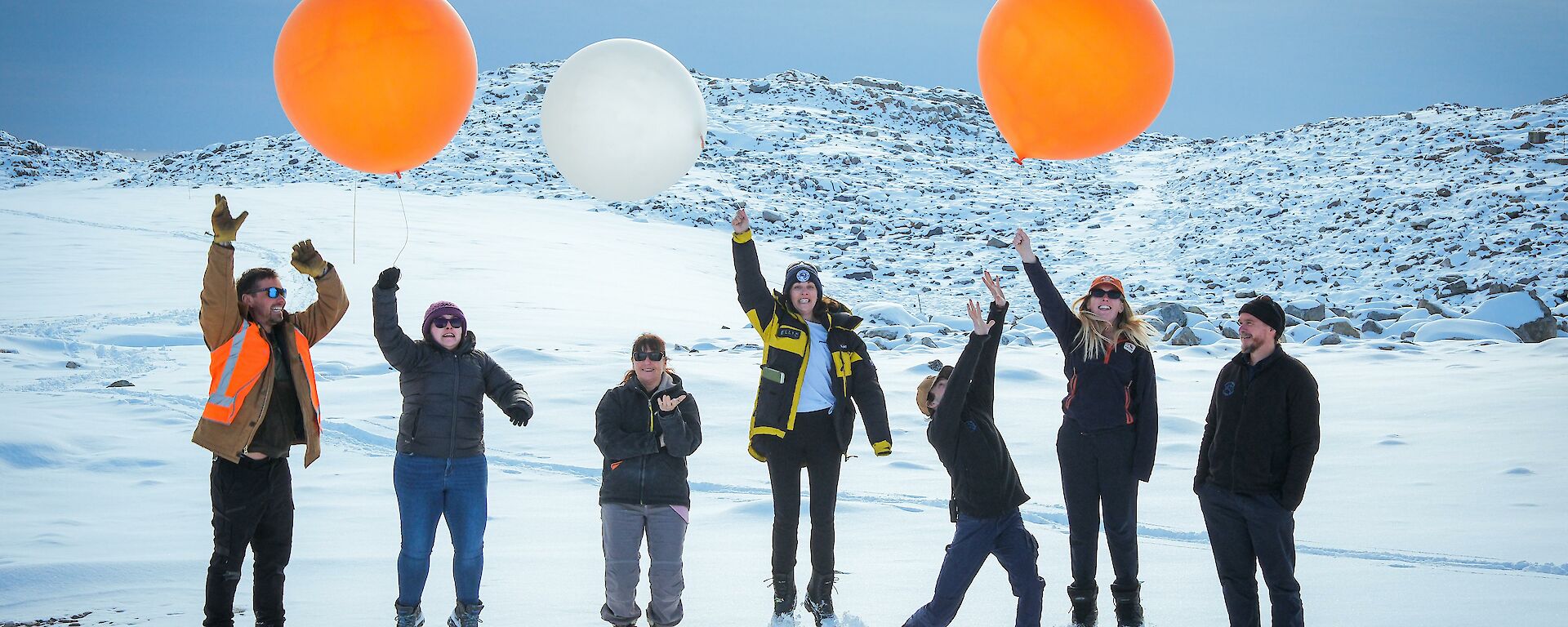 Seven expeditioners from the Bureau of Meteorology pose for a photo holding some weather balloons within Casey station a snowy hill pictured in background