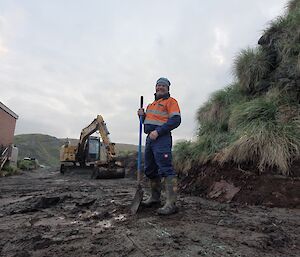 A man stands smiling on a muddy road. He has a shovel and there is an excavator in the background