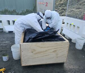 Two people are outside in hazmat suits putting a black back into a wooden box