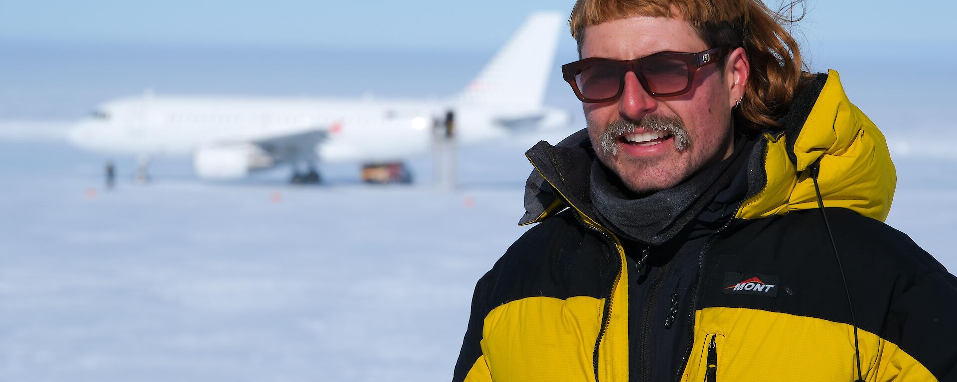 An expeditioner with wild hair poses for a photo at Wilkins aerodrome with an A319 plane in the backround