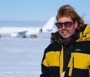 An expeditioner with wild hair poses for a photo at Wilkins aerodrome with an A319 plane in the backround