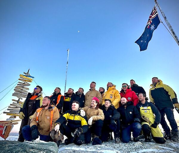 16 expeditioners pose under the Australian flag pole and Casey sign in support of a mental health initiative.