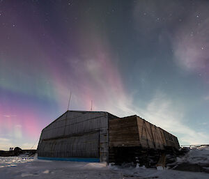 A rainbow of auroral lights in the night sky with the old hangar in the foreground near Mawson station on 25 March 2024