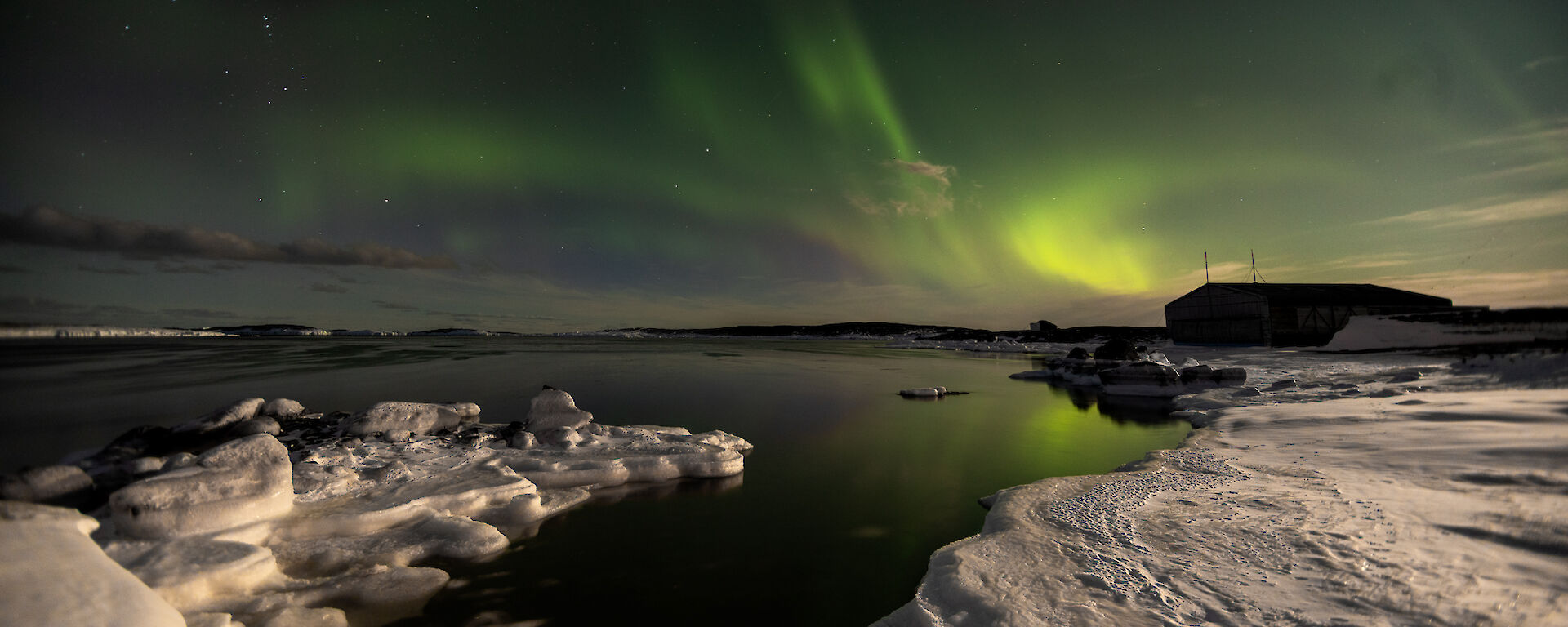 Green swirls of auroral lights mirrored in the perfectly calm ocean fringed with icy rocks near Mawson on 25 March 2024
