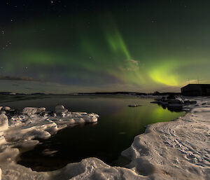 Green swirls of auroral lights mirrored in the perfectly calm ocean fringed with icy rocks near Mawson on 25 March 2024