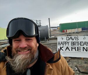 A man stands in front of a signboard with station buildings behind him saying 'Davis happy birthday Karen'