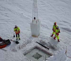 Three people in bright yellow stand on ice while a massive cube is cut out of it